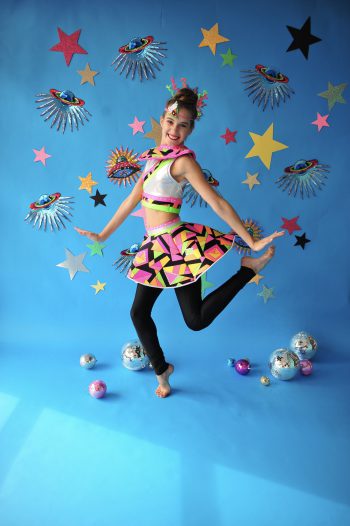 Teen dancer posing in a cute space themed costume in front of a blue background with stars and space ships.