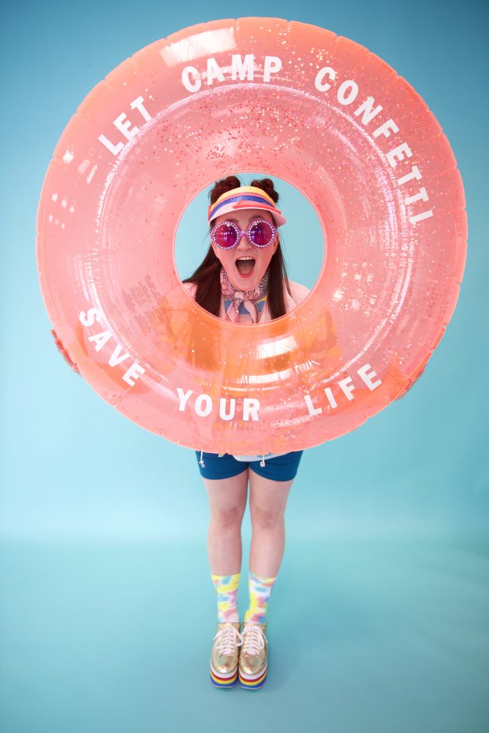 Happy dance teacher who is planning a summer dance camp is dressed in summer apparel holding life saver float that says Let Camp Confetti Save Your Life!