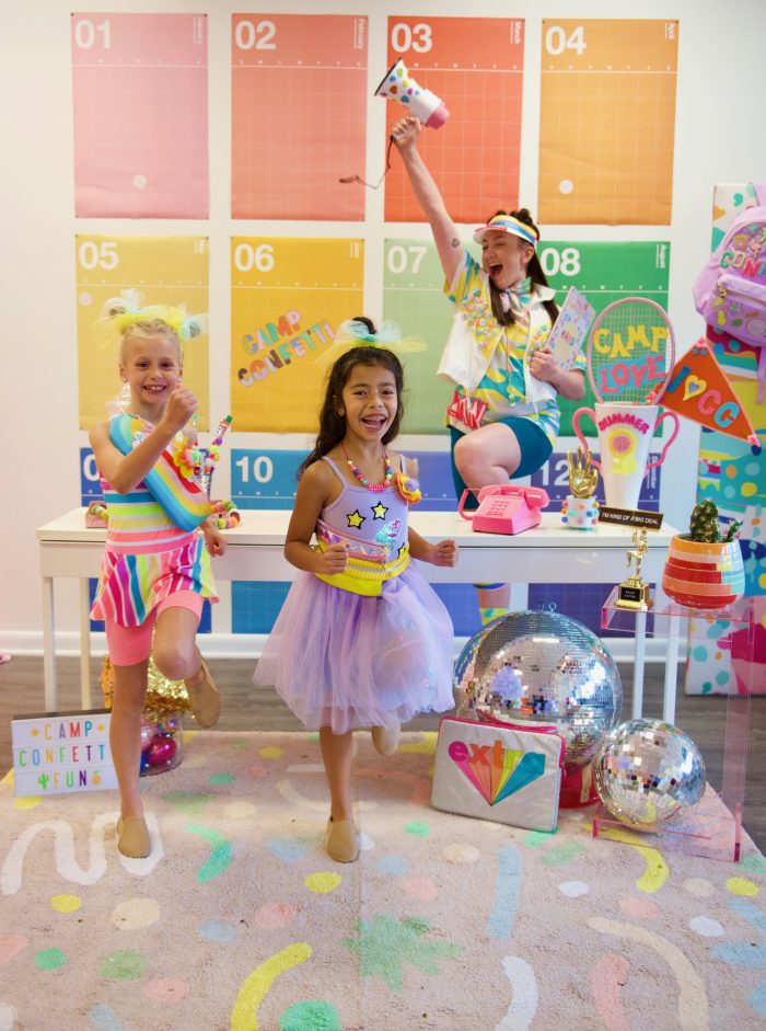 A dance teacher poses in front of colorful calendar back drop with excitement that her summer dance camps are planned for her through Camp Confetti. Two elementary-aged dancers are laughing, smiling and dancing in front of her. 