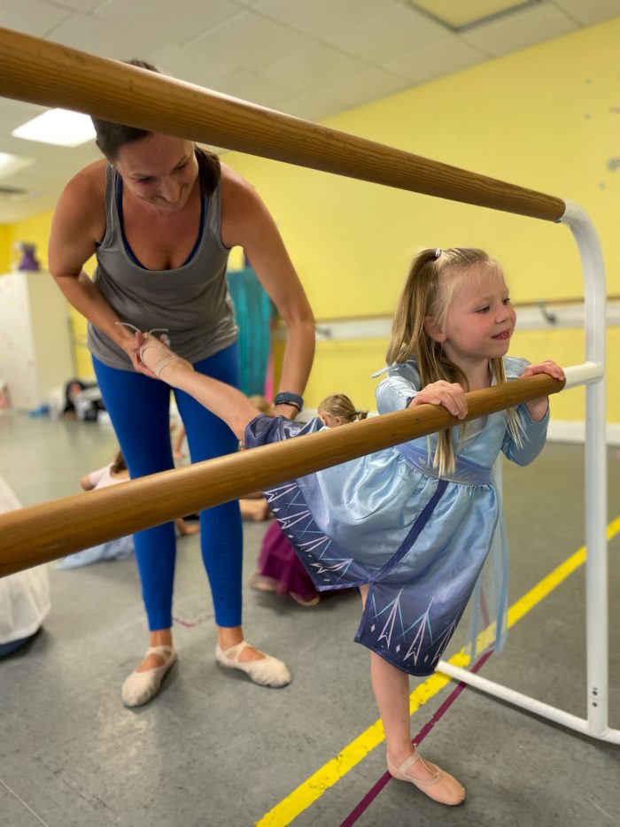 Dance teacher assists young dancer dressed as a princess at the ballet barre during a summer dance camp.