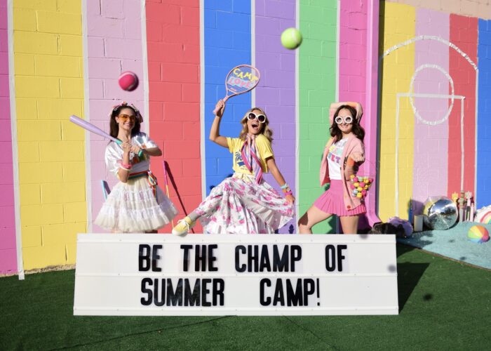 Dancers dressed in party style sports attire holding sports equipment in front of pastel rainbow wall with a sign that says Be The Champ of Summer Camp!