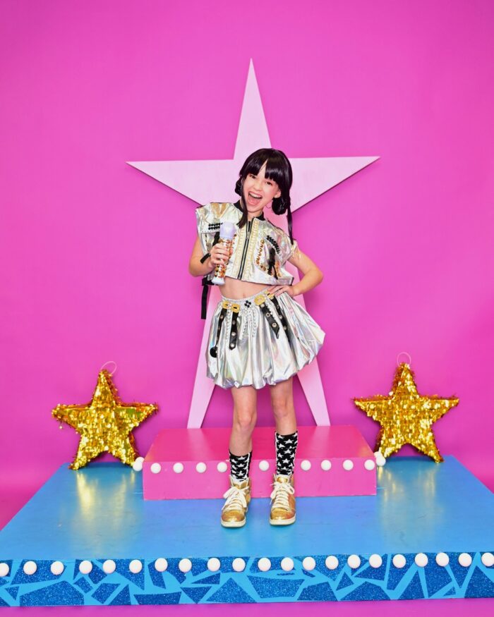 Dancer on stage holding a microphone in front of a pink background with star decorations.