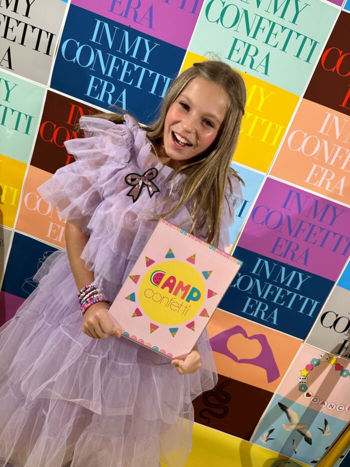 Swiftie dancer wearing a purple frilly dress standing in front of Taylor Swift Eras Tour inspired background holding a sign that says Camp Confetti