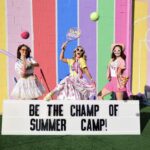 Dancers dressed in party style sports attire holding sports equipment in front of pastel rainbow wall with a sign that says Be The Champ of Summer Camp!