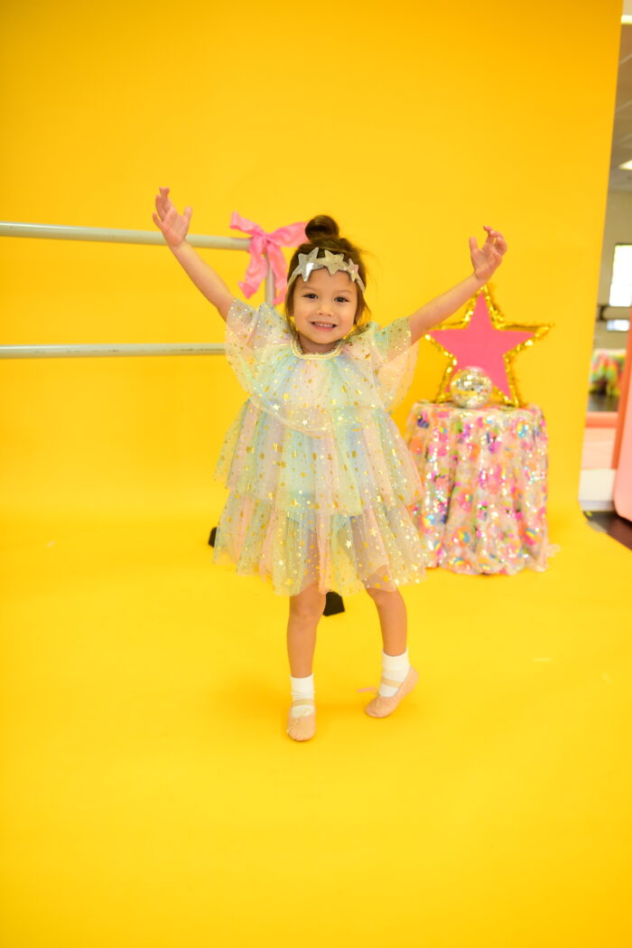 Preschool dancer wearing sparkly tutu and headband dancing in ballet shoes in front of a ballet barre on a yellow background.